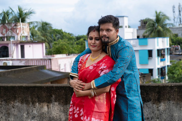 A brunette Indian Bengali romantic couple in traditional wear enjoying themselves standing on the roof top in sunny morning of Durga Puja festival in green natural background. Indian lifestyle.