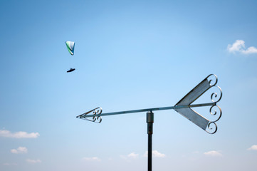 Arrow shape weathervane against summer's sky with cumulus and paragliders in background, italian Alps. Italy