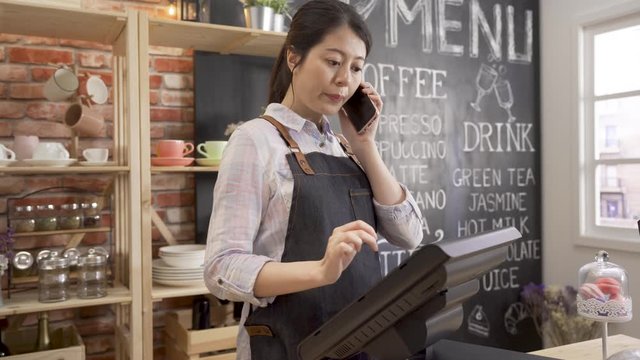 Female barista talking on mobile phone with customer and using tablet while standing behind counter in cafe store. young girl waitress in coffeehouse taking client order on cellphone call online.