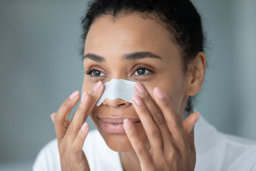 Close up face of 30s African ethnicity woman apply white cleansing nose strip for deep purifying and blackheads removal. Modern cosmetology treatment for ideal healthy beautiful skin, skincare concept