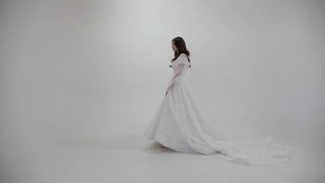 long haired brunette in white open shoulder wedding dress walks along white room under electric light slow motion