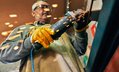 Bottom view horizontal image of adult worker man manufacturing in his workshop. Mature carpenter male grinding with sparks in repair shop.