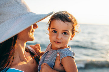 Closeup portrait of happy beautiful mother hugs her cute daughter, wearing white hat and striped dress posing at the sea. Mom with little girl walking at the ocean. Female with baby posing at ocean