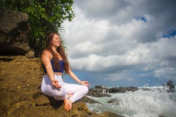 outdoors portrait of young beautiful and happy red hair woman practicing yoga and meditation exercise in lotus position at beach rock in healthy lifestyle and balance concept