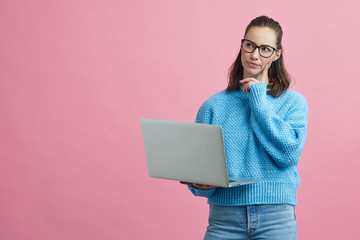 Smart looking student with glasses thinking and standing with her laptop. Colorful portrait concept