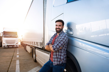 Portrait of young Caucasian bearded trucker with arms crossed standing by his truck vehicle. Transportation service. - obrazy, fototapety, plakaty
