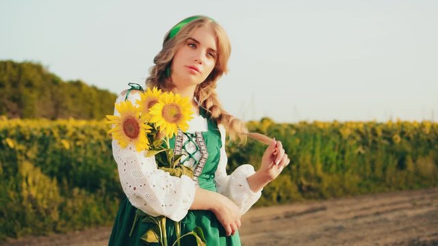 German authentic impartial young beautiful blonde woman holding bouquet yellow sunflowers. Green national traditional costume. Bare foot. Backdrop blue fabulous sky, flowering field. Harmony nature