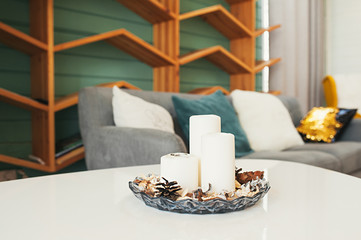 white candles in a candlestick for aromatherapy against the background of a cozy apartment with a gray sofa, cozy pillows and a wooden shelf for books