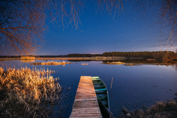 Belarus, Eastern Europe. Real Night Sky Stars Above Old Pier With Moored Wooden Fishing Boat. Natural Starry Sky And Countryside Landscape With Lake River In Early Spring Night. Russian Nature