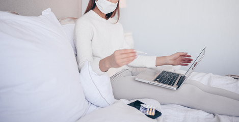 Unrecognizable sick woman in face protection mask lying in bed with laptop holding thermometer and pills at home quarantine isolation. Online work from home. Corona virus infection COVID-19 concept. 