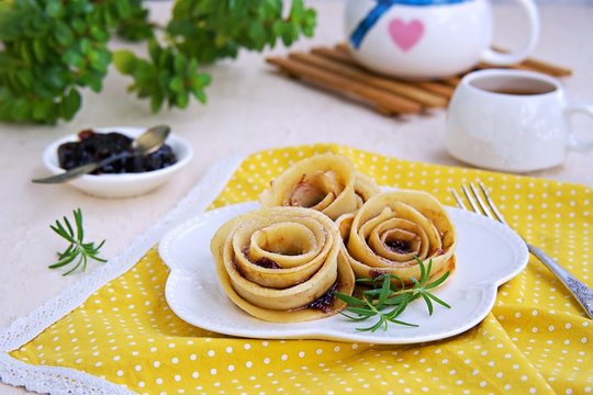 Pancakes, rolled up in the shape of a rose flower, on a white plate on a light concrete background. Pancake Day concept. Pancake Recipes. Served with plum jam