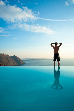 Unrecognizable man standing on the edge of an infinity pool looking out over a dramatic Mediterranean view of the Santorini caldera, Greece