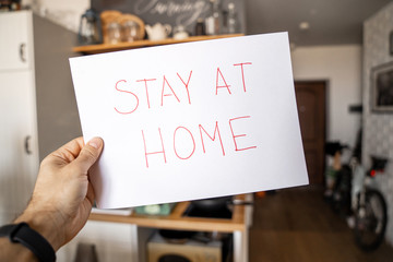 The inscription on a sheet of paper in the hands of a person "stay home", against the background of the interior of the apartment.