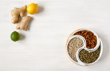 Useful plant-based vegetarian products. Sunflower seeds, pumpkin and raisins in a beautiful round plate divided into compartments, next to the roots of ginger lemon and lime, on a white wooden table.