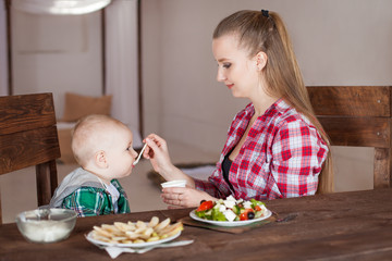 Beautiful woman in a red checkered shirt feeds her baby with a white spoon. A one-year-old child in a green shirt is trying to eat on his own, sitting on a brown wooden chair. The fair-haired girl 