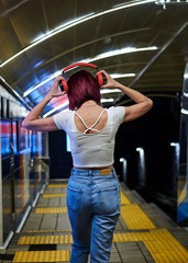 Young woman with pink burgundy maroon hair, wearing white top and light blue jeans, holding her red headphones on the head, walking in subway station, with her back towards the camera.
