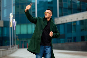 Young business man in green coat in business center with smartphone make selfie