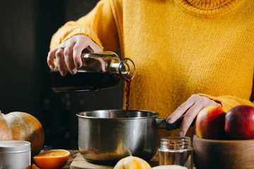 A young woman in a warm, knitted, yelow pullover is pouring wine from glass bottle to a pan to make hot mulled wine with spices and citrus fruits. A cozy, winter evening. Winter drinks.