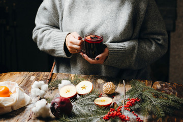 Hot mulled wine in the woman hands in grey sweater. Beautiful Christmas decorations lying on the table: fur-tree branches, red berries, apples, oranges, cotton flower. Festive mood.