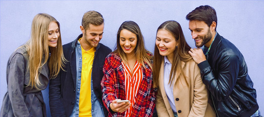 Young people standing by the wall while looking in smartphone and smiling- College friends checking...