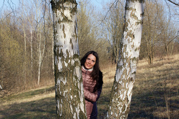 A girl walks through the spring park. Peeks between the trunks of birches.