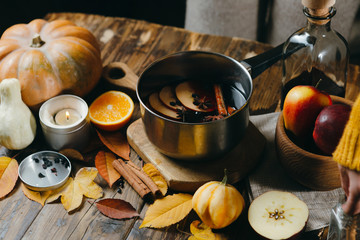 Mulled wine hot drink with citrus, apple and spices in aluminum pot on wooden background. Autumn mood with pumpkins, candles and leaves.