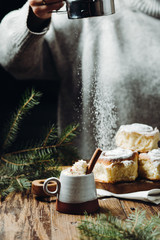 A hot cocoa with cinnamon stick and fresh Christmas buns with powder and over festive table, a girl is sprinkling sugar powder on the background. Winter holidays.