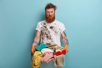Displeased dirty man with ginger beard stands with basin full of laundry, doesnt want to wash by hands, tired of housekeeping process, has dirty face and clothes, feels laziness during cleaning day