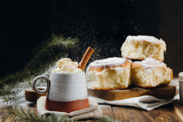 A hot cocoa with whipped cream, cinnamon stick and fresh Christmas buns with powder and over festive table, sugar powder is sprinkled on the background lke a snow. Winter holidays.
