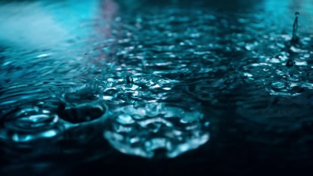 Slow motion of rain drops falling into puddle on asphalt at night. Big modern city lights mirror reflection on water surface. Close-up of raining outside in wet season
