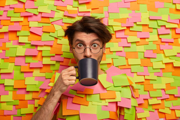 Surprised male face sips coffee or tea from cup, poses against creative background in office, surrounded by colorful stickers, wears round spectacles, listens interlocutor attentively, feels shock