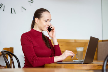 Young woman works and speaks on the phone with laptop and a coffee at home