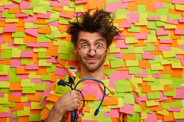 Funny repairman holds bunch of computer cables, poses with electronic wires, has messy hair wears...