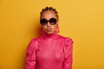 Beautiful cheeky woman keeps lips rounded, wears trendy big sunglasses and pink blouse, has glamour look, isolated on yellow background. Fashion model dressed in stylish outfit. Beauty trends