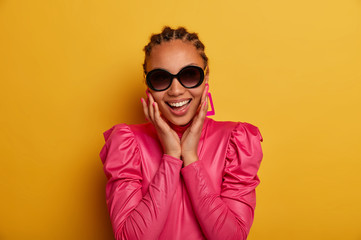 Gorgeous happy woman in fashion wear and trendy shades, keeps hands on face, has healthy skin, perfect teeth and manicure, smiles happily isolated on yellow background. Beauty portrait. Spring fashion