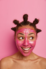 Close up shot of funny dark skinned woman applies glittering beauty mask on face, looks aside with toothy smile, stands bare shoulders, spends much time on skin care, isolated over pink background