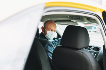 Bald man taxi driver in medical face mask inside yellow car looks at camera, concept of coronavirus...