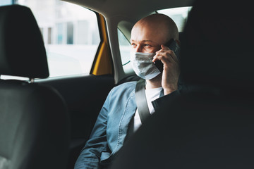 Bald man businessman in medical face mask using mobile phone inside yellow car taxi, Concept of...