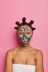 Beauty, wellness concept. Funny young female applies facial mask for healthy skin, keeps lips rounded, has knot hairstyle, poses with bare shoulders wrapped in towel, isolated on pink background