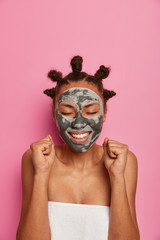 Photo of happy ethnic woman clenches fists, rejoices nice effect after applying beauty mask, has smooth perfect skin, smiles broadly and closes eyes with pleasure, isolated on pink background