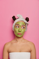 Young woman wears natural facial mask, looks aside thoughtfully, feels sad, has skin treatments after taking bath, poses indoor against pink background. Beauty, cosmetics, healthy lifestyle concept