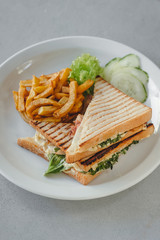 Bacon club sandwich with fries and vegetables on a white plate on a white table. Meal time.