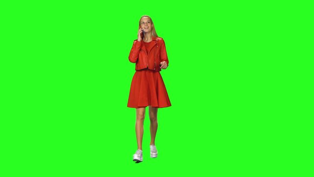 Blonde girl is calmly walking and talking on the mobile phone at green screen background. Chroma key. Front view.