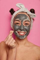 Spa treatment concept. Positive cheerful Asian woman smiles and makes korean like sign, looks at camera happily, stands naked, cares about facial skin, removes wrinkles, wears headband, poses indoor