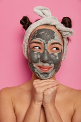 Beautiful young woman looks aside, has applied clay mask on face, enjoys beauty procedures in spa center, keeps hands under chin, waits for result of rejuvenation procedure, poses with naked body