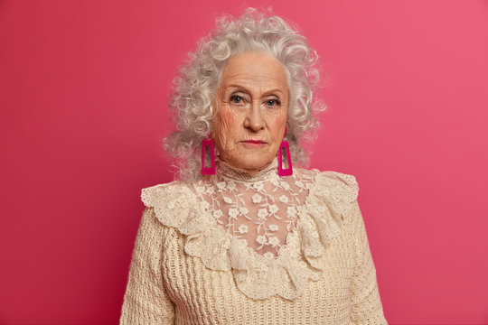 Portrait of beautiful curly senior lady looks seriously at camera, dressed in fashionable clothes, prepares for festive event, poses in studio against pink background, looks well in her old age