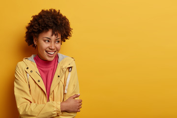 Obraz na płótnie Canvas Half length shot of positive Afro American woman looks happily and carefree aside, wears casual jacket, poses against yellow background, blank space, hears hilarious story, has friendly conversation