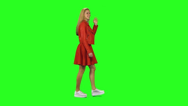 Blonde girl calmly walking and wave her hand in greeting on green screen background. Chroma key, 4k shot. Profile view.