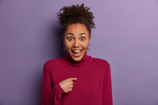 Happy excited ethnic woman points at herself, being told good news, feels satisfaction of achieving success, wears casual turtleneck, models against purple wall. Lucky winner cannot believe in victory