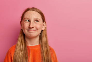 Funny freckled redhead girl looks happily aside, has tricky plan, smiles with cunny expression,...